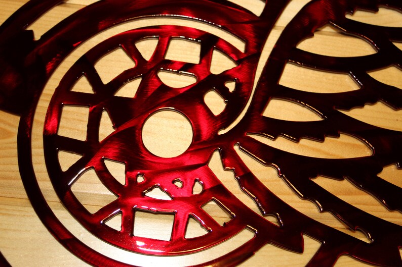 May include: A close-up of a red metal wall art piece with a circular design and intricate cut-out patterns. The artwork has a glossy finish and is mounted on a light-colored wooden surface. The design features a central circle and wing-like shapes.