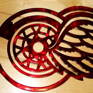 May include: A red metal wall art piece featuring a stylized wing and a circular emblem. The design has a glossy finish and is likely a sports team logo. The artwork is mounted on a light-colored wooden surface.