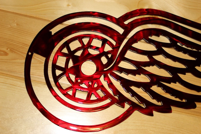 May include: A red metal wall art piece featuring a stylized wing and a circular emblem. The design has a glossy finish and is likely a sports team logo. The artwork is mounted on a light-colored wooden surface.