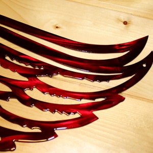 May include: A close-up of a red metal wing sculpture with a glossy finish. The wing design features curved lines and jagged edges, mounted on a light-colored wooden surface. The artwork is a decorative piece.