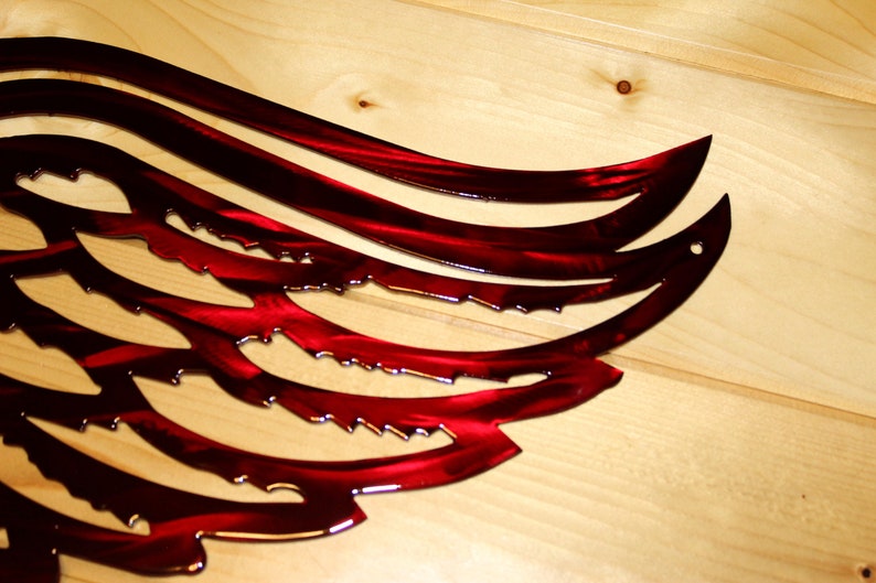 May include: A close-up of a red metal wing sculpture with a glossy finish. The wing design features curved lines and jagged edges, mounted on a light-colored wooden surface. The artwork is a decorative piece.