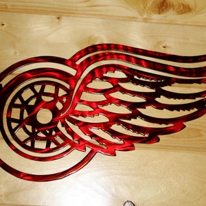 May include: A red metal wall art piece featuring the Detroit Red Wings logo, a winged wheel. The artwork is cut from metal and has a textured finish. The logo is mounted on a light-colored wooden surface.