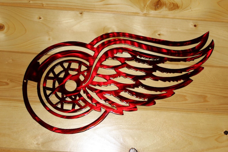 May include: A red metal wall art piece featuring the Detroit Red Wings logo, a winged wheel. The artwork is cut from metal and has a textured finish. The logo is mounted on a light-colored wooden surface.