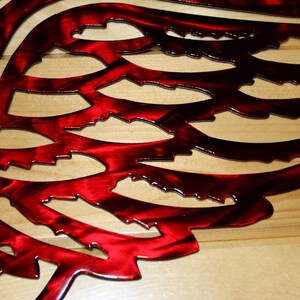 May include: A close-up of a red metal wing sculpture with intricate cut-out details. The wing is a vibrant red with a metallic sheen, and the design is mounted on a light-colored wooden surface. The artwork is a decorative wall hanging.