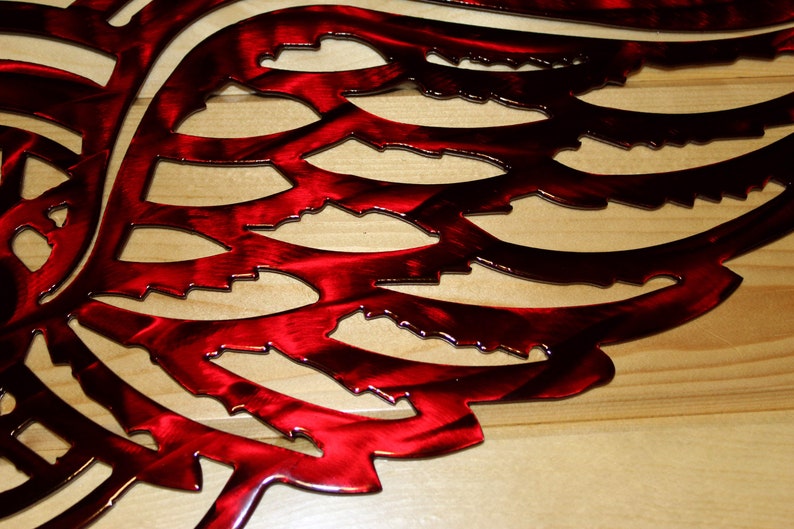 May include: A close-up of a red metal wing sculpture with intricate cut-out details. The wing is a vibrant red with a metallic sheen, and the design is mounted on a light-colored wooden surface. The artwork is a decorative wall hanging.