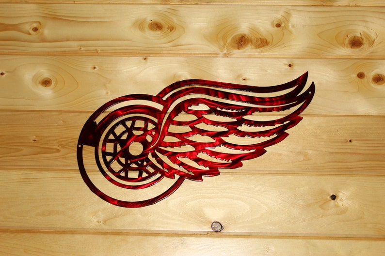 May include: A red and black metal Detroit Red Wings logo, featuring a wheel and wing design. The logo is mounted on a light brown wooden background, showcasing the team's emblem.