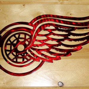 May include: A red metal Detroit Red Wings logo, a stylized winged wheel, is mounted on a light wood background. The logo is cut from metal, with a textured finish, and is designed to be mounted on a wall.
