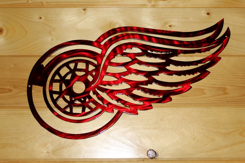 May include: A red metal Detroit Red Wings logo, a stylized winged wheel, is mounted on a light wood background. The logo is cut from metal, with a textured finish, and is designed to be mounted on a wall.