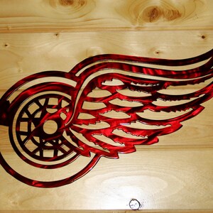 May include: Red and silver metal wall art featuring the Detroit Red Wings logo. The logo is a stylized wheel with a wing design, cut from metal. The art is mounted on a light-colored wooden background.