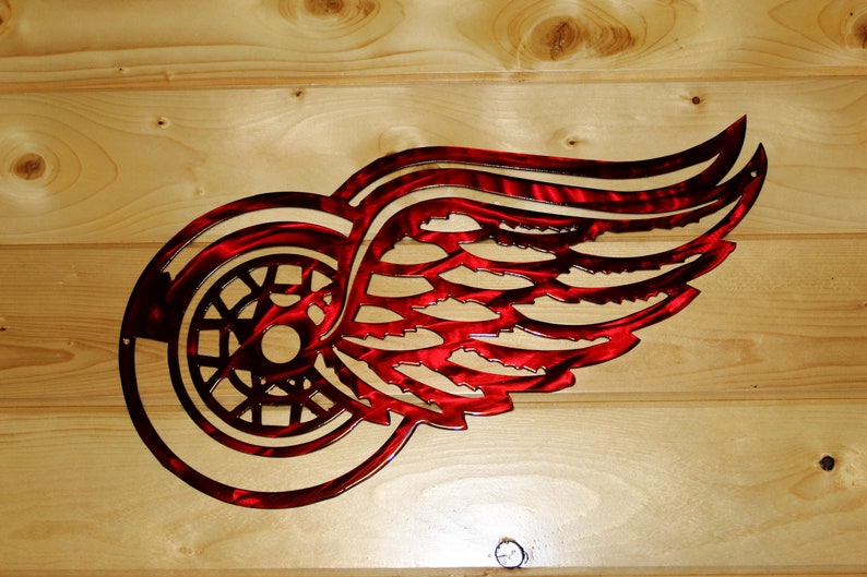May include: Red and silver metal wall art featuring the Detroit Red Wings logo. The logo is a stylized wheel with a wing design, cut from metal. The art is mounted on a light-colored wooden background.