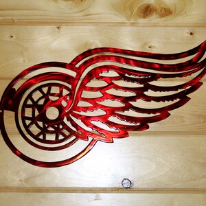 May include: A red metal wall art piece featuring the Detroit Red Wings logo, a stylized winged wheel. The artwork is mounted on a light-colored wooden background, showcasing the team's emblem.