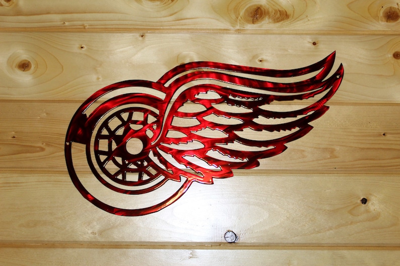 May include: A red metal wall art piece featuring the Detroit Red Wings logo, a stylized winged wheel. The artwork is mounted on a light-colored wooden background, showcasing the team's emblem.