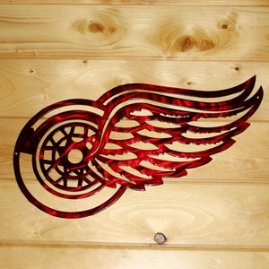 May include: Red and black metal wall art featuring the Detroit Red Wings logo, a winged wheel. The artwork is mounted on a light-colored wooden plank background. The metal has a textured finish.