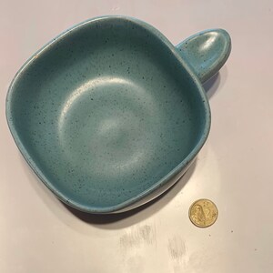 May include: A speckled, pale blue ceramic bowl with a handle. The square bowl has rounded corners and a smooth interior. The handle is a curved extension of the rim. A coin is next to the bowl.