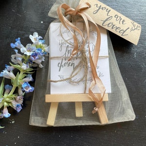 May include: A small wooden easel with a white card that reads "You are dearly loved & chosen" and a brown tag that reads "You are loved". The easel is wrapped in a sheer beige bag with a brown ribbon.