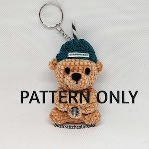 Pode incluir: Um chaveiro de urso de pelúcia de crochê em tons de marrom, usando um chapéu verde com o logotipo da Starbucks. O urso tem uma palhinha pequena e um chaveiro prateado. O texto "PATTERN ONLY" é exibido na imagem.