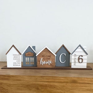 May include: A set of five wooden house-shaped ornaments with different designs. The first house is white with the names Manuel, Jessica, Damian, Liam, Aislynn, and Rosalia written on it. The second house is gray with the number 24653 on it. The third house is brown with the text "Welcome to our home" on it. The fourth house is gray with the letter "C" on it. The fifth house is white with the text "Party of 6" on it.