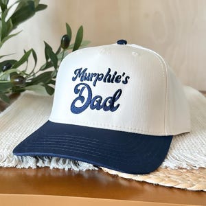 May include: A white baseball cap with a navy blue brim. The cap features the embroidered text "Murphie's Dad" in navy blue script. The hat is displayed on a wooden surface with a decorative woven mat and greenery in the background.