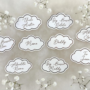 May include: White cloud-shaped place cards with gold lettering. The names on the cards include "Uncle Austin", "Big Cousin Natalie", "Big Cousin Alan", "Abuelito Elpidio", "Mama", "Daddy", "Tio Juan", "Abuelita Norma", and "Big Cousin Maddy".