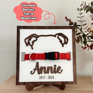 May include: A wooden plaque with a cutout of a dog's head, a red collar, and the name "Annie" with the dates 2017-2024. The plaque is on a small wooden stand.