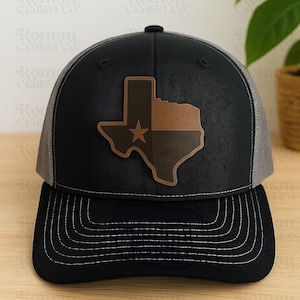 May include: Black and gray trucker hat featuring a leather patch of the Texas state outline with a star. The hat has a curved brim and a mesh back. The text "Roman Crafted Co." is visible in the background.