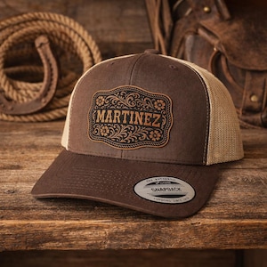 Może przedstawiać: Brązowo-beżowa czapka typu snapback z ozdobną skórzaną naszywką. Na naszywce widnieje imię "MARTINEZ" w stylu western, otoczone kwiatowymi i przewijanymi wzorami. Czapka ma zakrzywiony daszek i siateczkowy tył.