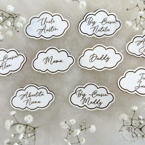 May include: White cloud-shaped place cards with gold trim and calligraphy names, including "Uncle Austin", "Big Cousin Natalie", "Cousin Alan", "Mama", "Daddy", "Abuelita Norma", "Big Cousin Maddy", and "Ju".