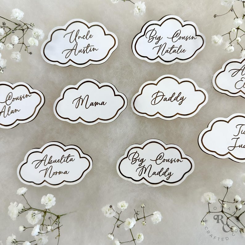 May include: White cloud-shaped place cards with gold trim and calligraphy names, including "Uncle Austin", "Big Cousin Natalie", "Cousin Alan", "Mama", "Daddy", "Abuelita Norma", "Big Cousin Maddy", and "Ju".