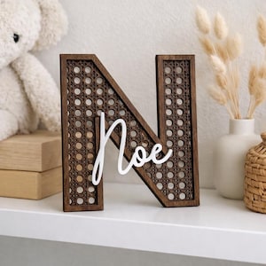May include: A wooden letter "N" with a woven cane design, featuring the name "Noe" in white script. The dark brown letter stands on a white surface, suggesting a decorative item for a nursery or home decor.