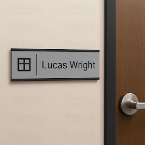 May include: A rectangular office door sign with the name "Lucas Wright" in black text. The sign has a gray background with a black border and a graphic of a house. The sign is mounted on a beige wall.