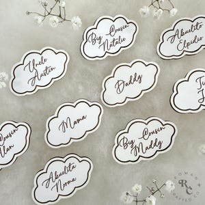 May include: White cloud-shaped place cards with brown calligraphy names, including "Uncle Austin", "Mama", "Daddy", "Big Cousin Natalie", "Big Cousin Maddy", "Abuelita Norma", "Abuelito Elpidio", and "Cousin Ilan".