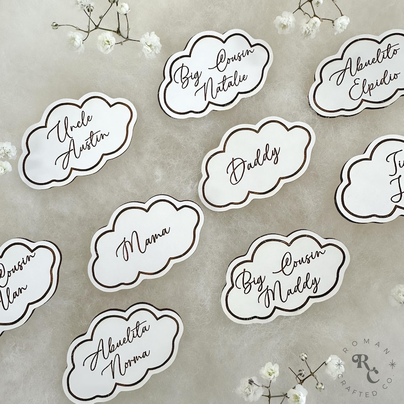 May include: White cloud-shaped place cards with brown calligraphy names, including "Uncle Austin", "Mama", "Daddy", "Big Cousin Natalie", "Big Cousin Maddy", "Abuelita Norma", "Abuelito Elpidio", and "Cousin Ilan".