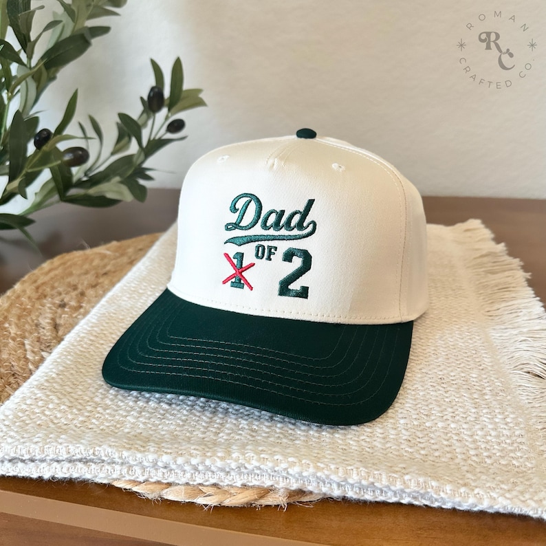 Puede incluir: Gorra de b&eacute;isbol blanca y verde con el texto bordado "Dad of 2". La gorra tiene una visera verde oscuro y un bot&oacute;n negro. El sombrero est&aacute; sobre una superficie blanca texturizada.