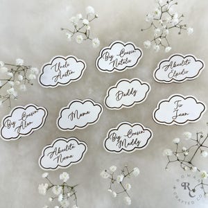 May include: White cloud-shaped place cards with gold trim and black calligraphy. The cards feature names like "Uncle Austin", "Big Cousin Alan", "Mama", "Abuelita Norma", "Big Cousin Natalie", "Daddy", "Big Cousin Maddy", "Abuelito Elpidio", and "Tio Juan".