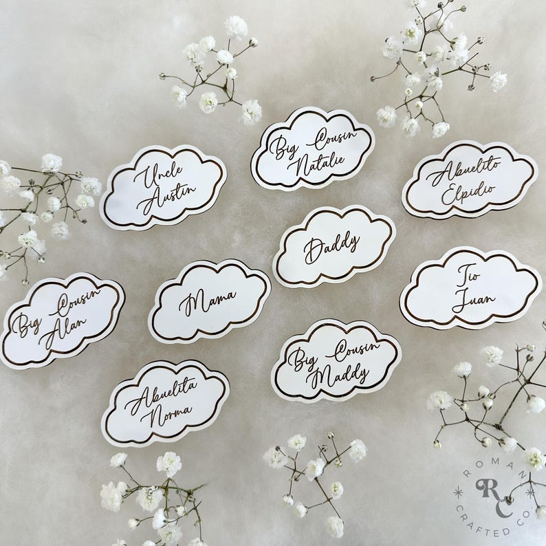 May include: White cloud-shaped place cards with gold trim and black calligraphy. The cards feature names like "Uncle Austin", "Big Cousin Alan", "Mama", "Abuelita Norma", "Big Cousin Natalie", "Daddy", "Big Cousin Maddy", "Abuelito Elpidio", and "Tio Juan".