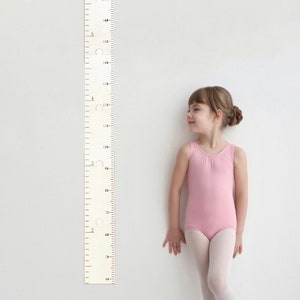 Pode incluir: Um gr&aacute;fico de crescimento de madeira branco com marcas pretas e um adesivo roxo e branco com o texto "Camila & Mia". Uma menina com um collant rosa e meias brancas fica ao lado do gr&aacute;fico.
