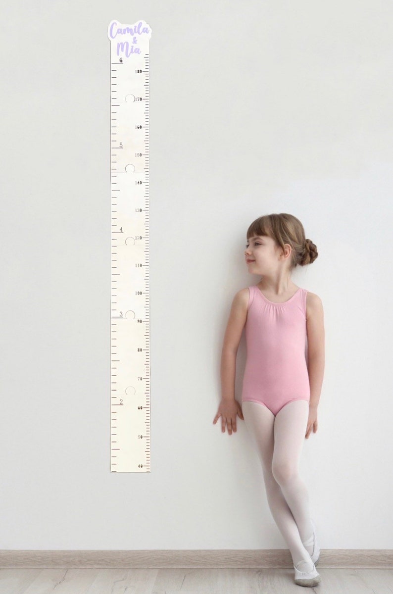 Pode incluir: Um gr&aacute;fico de crescimento de madeira branco com marcas pretas e um adesivo roxo e branco com o texto "Camila & Mia". Uma menina com um collant rosa e meias brancas fica ao lado do gr&aacute;fico.
