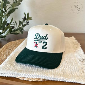 Puede incluir: Gorra de b&eacute;isbol crema y verde oscuro con el texto bordado "Dad of 2". La gorra presenta una visera verde oscuro y un peque&ntilde;o bot&oacute;n verde. El sombrero se muestra sobre una superficie tejida blanca.