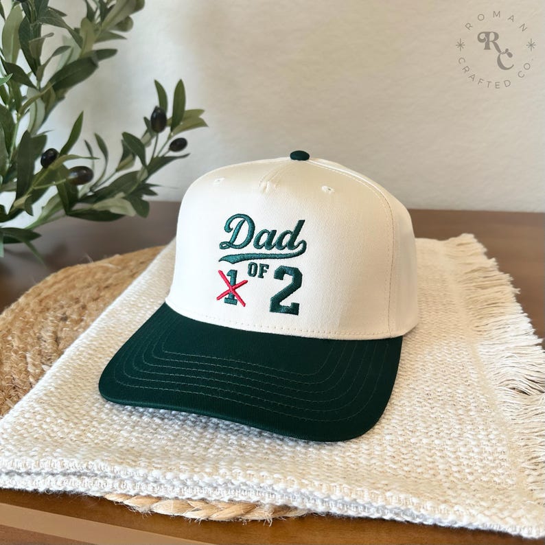 Puede incluir: Gorra de b&eacute;isbol crema y verde oscuro con el texto bordado "Dad of 2". La gorra presenta una visera verde oscuro y un peque&ntilde;o bot&oacute;n verde. El sombrero se muestra sobre una superficie tejida blanca.