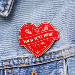Puede incluir: Un pin rojo en forma de corazón con un borde blanco y una pancarta que dice "YOUR TEXT HERE". El pin presenta diseños florales y de flechas. Está prendido a una chaqueta vaquera azul.