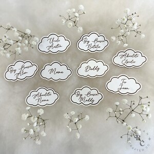 May include: White cloud-shaped place cards with black outlines and brown calligraphy. The cards feature names like "Uncle Austin", "Big Cousin Natalie", "Big Cousin Alan", "Abuelito Elpidio", "Mama", "Daddy", "To Juan", "Abuelita Norma", and "Big Cousin Maddy".