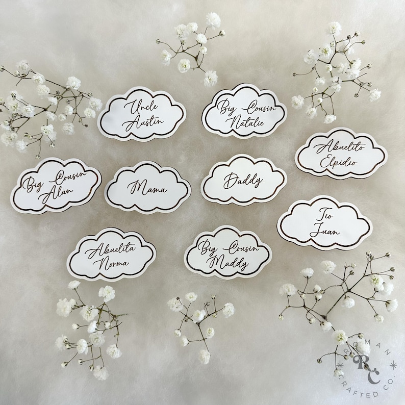May include: White cloud-shaped place cards with black outlines and brown calligraphy. The cards feature names like "Uncle Austin", "Big Cousin Natalie", "Big Cousin Alan", "Abuelito Elpidio", "Mama", "Daddy", "To Juan", "Abuelita Norma", and "Big Cousin Maddy".