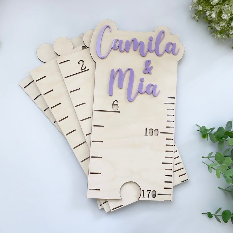 Pode incluir: Um gr&aacute;fico de crescimento de madeira com um recorte roxo dos nomes "Camila & Mia" e medidas em cent&iacute;metros. O gr&aacute;fico est&aacute; empilhado sobre si mesmo, mostrando as m&uacute;ltiplas camadas.
