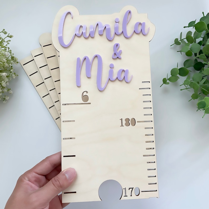 Pode incluir: Um gr&aacute;fico de crescimento de madeira com uma placa de nome de acr&iacute;lico roxo que diz "Camila & Mia". O gr&aacute;fico tem uma escala de 6 a 180 cent&iacute;metros.