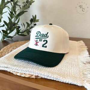 Puede incluir: Gorra de b&eacute;isbol crema y verde oscuro con la inscripci&oacute;n bordada "Dad of 2". La gorra tiene una visera verde oscuro y un peque&ntilde;o bot&oacute;n verde. Descansa sobre una superficie blanca texturizada, de estilo informal.