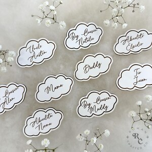 May include: White cloud-shaped place cards with gold lettering. The names on the cards include "Uncle Austin", "Mama", "Daddy", "Big Cousin Natalie", "Big Cousin Maddy", "Cousin Alan", "Abuelita Norma", "Abuelito Elpidio", and "Tio Juan".