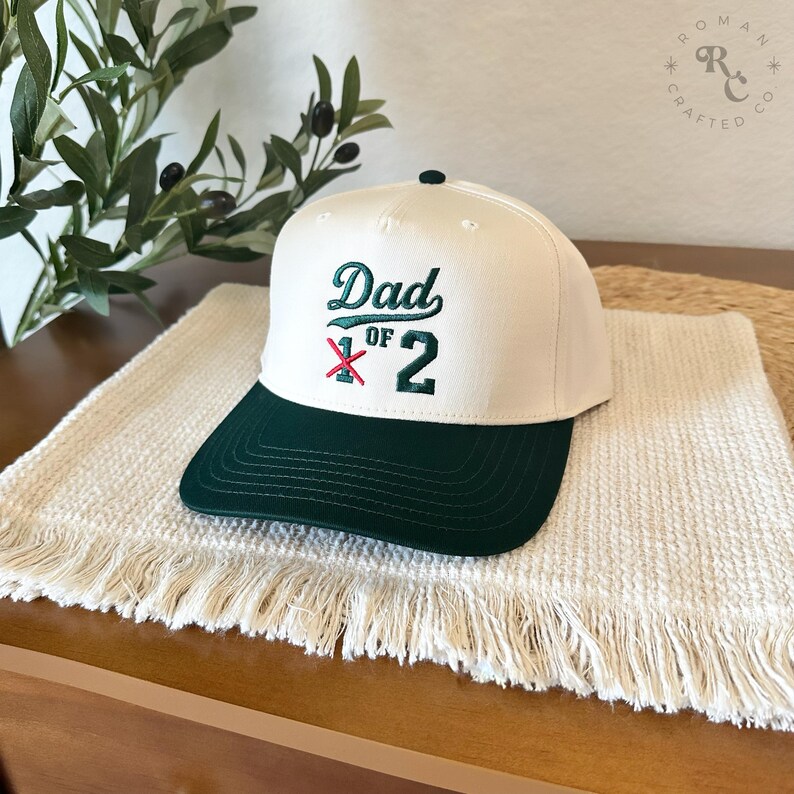 Puede incluir: Gorra de b&eacute;isbol blanca con visera verde oscuro. La gorra presenta el texto bordado "Dad of 2" en verde y rojo. El sombrero se exhibe sobre una esterilla blanca con flecos.