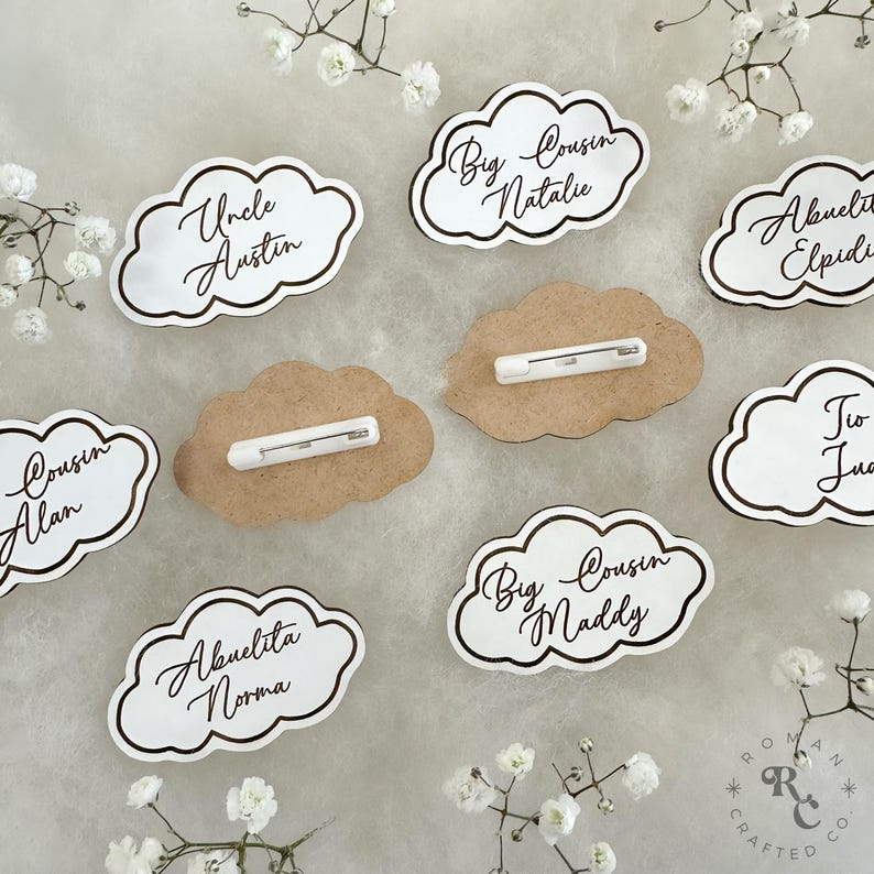 May include: A set of ten white cloud-shaped name tags with brown lettering. The name tags have a pin on the back for attaching to clothing. The names on the tags include "Uncle Austin", "Big Cousin Natalie", "Cousin Alan", "Abuelita Norma", "Big Cousin Maddy", "Abuelita Elpidia", "Tio Juan", and "Big Cousin Natalie".