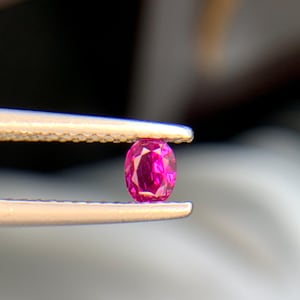 Puede incluir: Una pequeña piedra preciosa de corte ovalado con un color rojo rosado intenso y vibrante, con múltiples facetas que reflejan la luz. La piedra preciosa se sostiene de forma segura entre las dos puntas metálicas de unas pinzas.