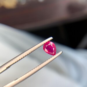 May include: A pear-shaped, faceted gemstone with a vibrant, deep red color. The stone is held by a pair of metal tweezers with a serrated grip, showcasing the clarity and precise cut of the gem against a soft, out-of-focus background.